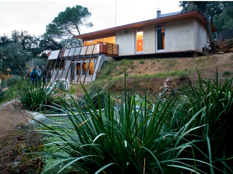 A modern house with large windows and a wooden deck, situated on a hillside surrounded by greenery and a small pond with lush plants in the foreground. Selfbuilding, Straw Bale, 